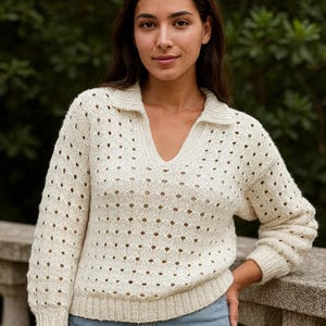 May include: Cream-colored, open-knit sweater with a collared V-neck. The long-sleeved top has a textured pattern with small holes throughout. The sweater is paired with light blue jeans. The model is leaning on a stone railing.