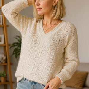 以下が含まれることがあります： 繊細な透かし模様が施された、クリーム色のVネックセーター。長袖ニットは、ライトブルーのデニムジーンズと組み合わされています。ゴールドのネックレスがさりげないアクセントに。セーターの質感とデザインが、居心地の良いスタイリッシュな外観を作り出しています。