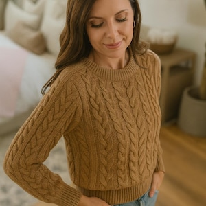 May include: A woman in a tan cable-knit jumper and blue jeans. The jumper features a crew neck and long sleeves with ribbed cuffs. The cable-knit pattern is prominent across the front and sleeves. The woman is indoors with her hands in her pockets.