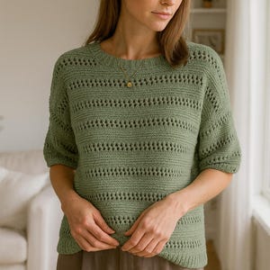 Peut inclure: Un pull en tricot vert sauge avec un col rond et des manches courtes. Le pull présente un motif ajouré horizontal. Le modèle porte un collier en or et un pantalon marron. Le pull est un vêtement confortable et élégant.