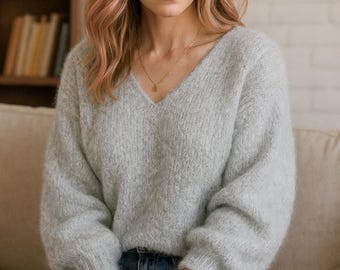 以下が含まれることがあります： ライトグレーのVネックセーター、長袖。セーターはブルージーンズと組み合わせています。ゴールドのネックレスと、明るい色のバンドが付いたスマートウォッチも表示されています。