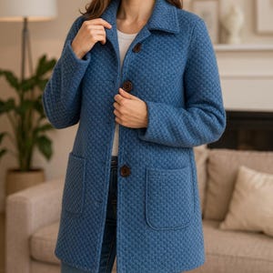 Op de afbeelding: Een blauwe jas met textuur met een reverskraag, twee voorzakken en drie bruine knopen. De jas is knielang en lijkt gemaakt van een geweven stof. Het model draagt een blauwe spijkerbroek en een wit shirt.