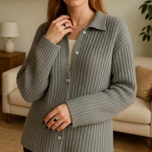 May include: A gray ribbed cardigan with a collared neckline and button-down front. The cardigan is worn over a white top. A silver necklace and a black ring are also visible. The background includes a sofa, a lamp, and a shelf.
