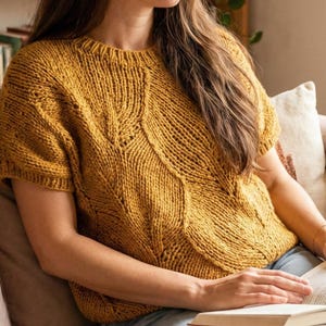 May include: A woman wearing a mustard yellow short-sleeved knit sweater with a textured design, sitting on a brown sofa and reading a book. The background includes a bookshelf, plants, and a window.