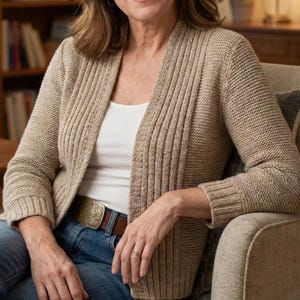 Könnte beinhalten: Eine Frau trägt einen beigefarbenen Cardigan über einem weißen Oberteil und einer blauen Jeans. Der Cardigan hat ein geripptes Design. Sie sitzt in einem beigen Sessel in einem Wohnzimmer.