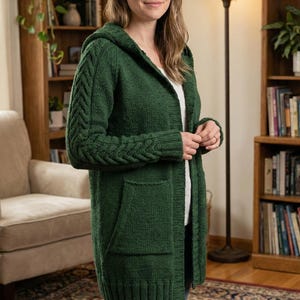 May include: A woman wearing a forest green hooded cardigan. The long-sleeved sweater has a cable knit design on the sleeves and two front pockets. The cardigan is knee-length and appears to be made of a soft, knitted material.