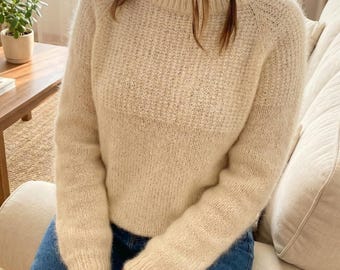 以下が含まれることがあります： クリーム色のクルーネックセーター、テクスチャードニットパターン。長袖セーターは柔らかくふわふわした素材で作られています。画像内の人物はブルージーンズを着用しています。セーターは居心地の良いカジュアルな衣服です。