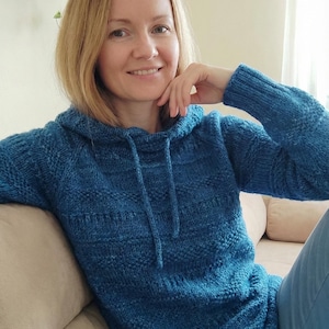 May include: A blue knitted hoodie with a drawstring, featuring a textured pattern. The long-sleeved sweater is a vibrant shade of blue. The model is wearing blue jeans. The hoodie is made of a soft, comfortable material.