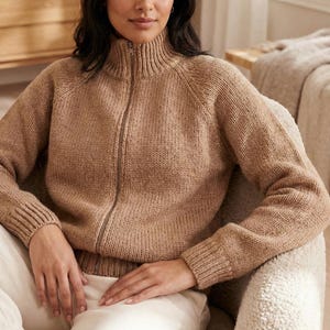 Peut inclure: Un pull zippé beige en tricot avec un col montant. Le pull a une fermeture éclair sur toute la longueur et des poignets et un ourlet côtelés. La personne est assise dans un fauteuil. Le pull est de couleur neutre.