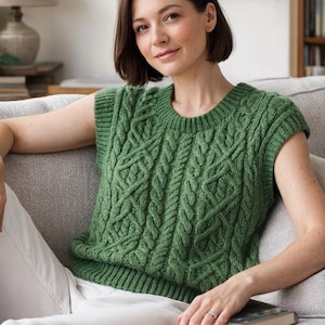May include: A green cable-knit sleeveless sweater with a round neckline. The sweater features a textured pattern and is worn with white pants. The person is sitting on a gray couch with pillows, holding a book.