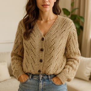 May include: A tan, open-knit cardigan with a V-neck and button closure. The long-sleeved sweater has a diamond pattern and is paired with blue jeans. The model is standing in front of a neutral background.