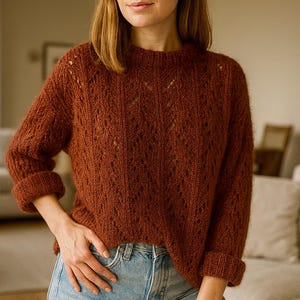 Puede incluir: Un suéter de punto color óxido con un patrón de diamantes texturizado. El suéter tiene mangas largas con puños enrollados y cuello redondo. La persona de la imagen lleva vaqueros azul claro.