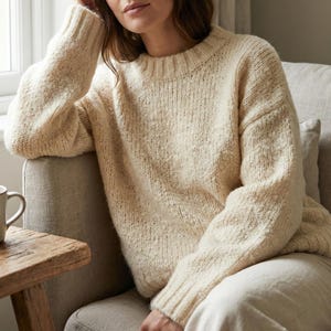 Peut inclure: Pull en tricot crème à col rond et manches longues. Le pull a un aspect texturé et est porté par une personne assise dans une chaise. Le pull est de couleur neutre et semble doux et confortable.
