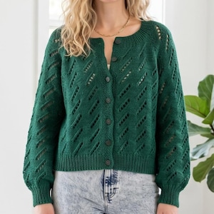 May include: A dark green, button-up cardigan with a round neck and long sleeves. The knitted jumper has a diamond-shaped openwork design. The sleeves are slightly puffed at the shoulders and cuffed at the wrists. The cardigan is made of a soft, textured yarn.