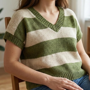 Puede incluir: Un suéter de cuello en V de manga corta con rayas horizontales en verde oliva y crema. La prenda de punto tiene un dobladillo y puños acanalados. La persona lleva vaqueros azules. El suéter es una prenda informal y cómoda.