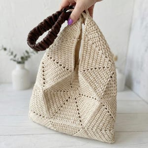 ALT TEXT (5)
1) Two-tone crochet tote bag in sage green and cream with a padded dark handle displayed on a light tabletop.
2) Side view of the patchwork crochet handbag showing triangle panels and a structured shape.
3) Close-up of the geometric triangle motif and textured stitches on the crochet tote bag.
4) Detail of the padded handle and hardware attachment at the top of the crochet bag.
5) Front view of the crochet tote highlighting the centered patchwork design and neat edging.