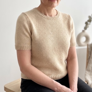 May include: A short-sleeved, beige knit sweater featuring a classic crew neckline. The garment is constructed with a smooth stockinette stitch pattern, complemented by ribbed detailing along the collar, sleeve cuffs, and the bottom hemline.
