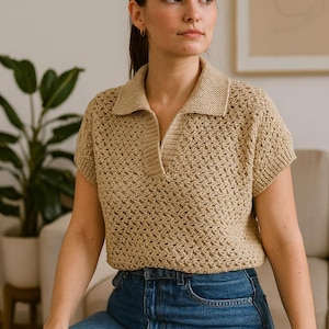 May include: A tan crochet polo shirt with short sleeves and a collared neckline. The shirt has a textured, open weave design. The person is wearing blue jeans. The background includes a plant and a neutral-toned wall.