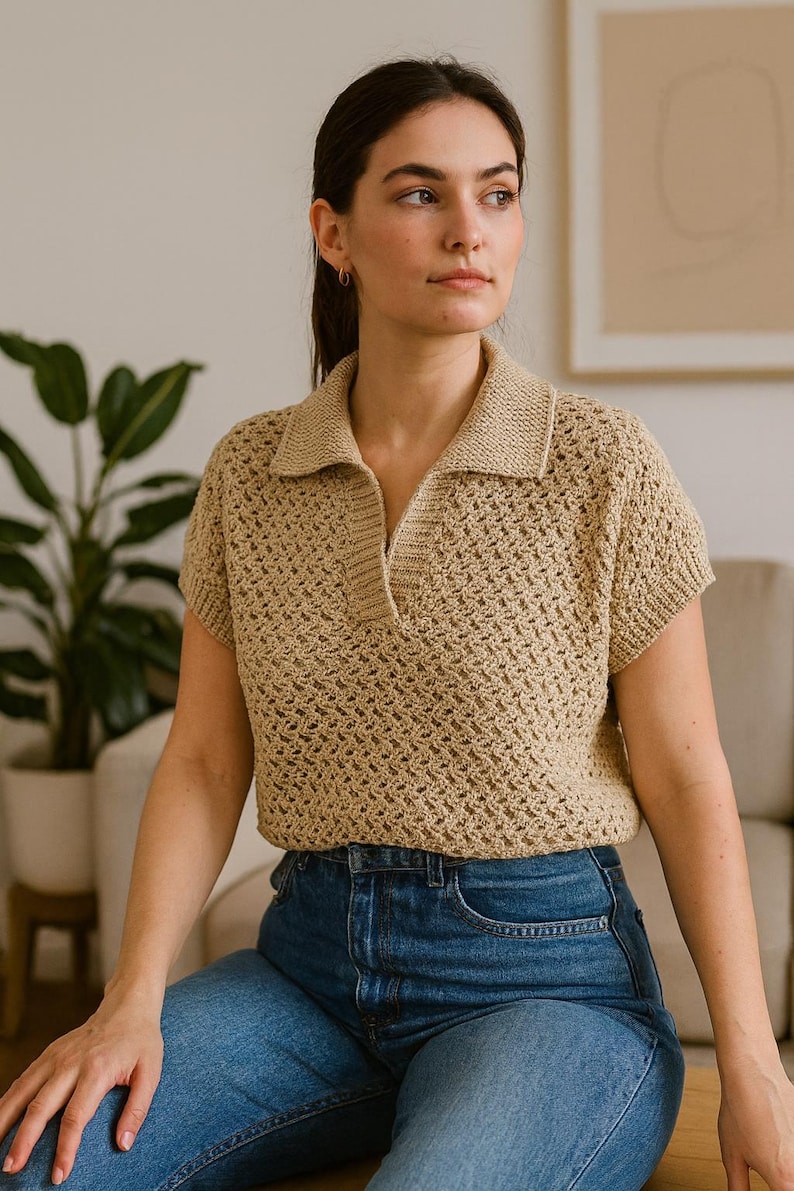 May include: A tan crochet polo shirt with short sleeves and a collared neckline. The shirt has a textured, open weave design. The person is wearing blue jeans. The background includes a plant and a neutral-toned wall.