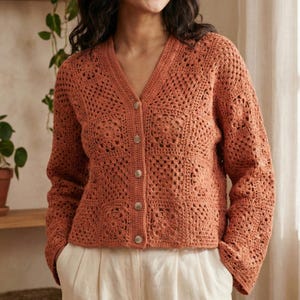 Può includere: Un cardigan all'uncinetto color ruggine con scollo a V e chiusura con bottoni. Il design a trama aperta presenta motivi floreali e geometrici. Il maglione a maniche lunghe è abbinato a pantaloni color crema.