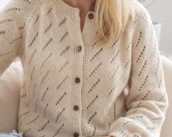 May include: A cream-colored long-sleeve knit cardigan featuring a repeating diagonal eyelet pattern, a ribbed crew neckline, and a front button closure with five small brown buttons. The garment is constructed with a soft, textured yarn.