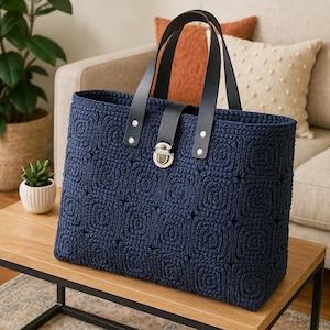 Może przedstawiać: Granatowa torba typu tote z szydełkowym wzorem, z czarnymi skórzanymi uchwytami i srebrnym zapięciem. Torba ma kwadratowy wzór i stoi na drewnianym stole. Stylowy dodatek.