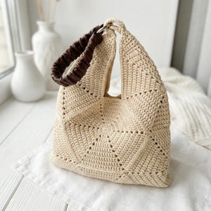 ALT TEXT (5)
1) Two-tone crochet tote bag in sage green and cream with a padded dark handle displayed on a light tabletop.
2) Side view of the patchwork crochet handbag showing triangle panels and a structured shape.
3) Close-up of the geometric triangle motif and textured stitches on the crochet tote bag.
4) Detail of the padded handle and hardware attachment at the top of the crochet bag.
5) Front view of the crochet tote highlighting the centered patchwork design and neat edging.