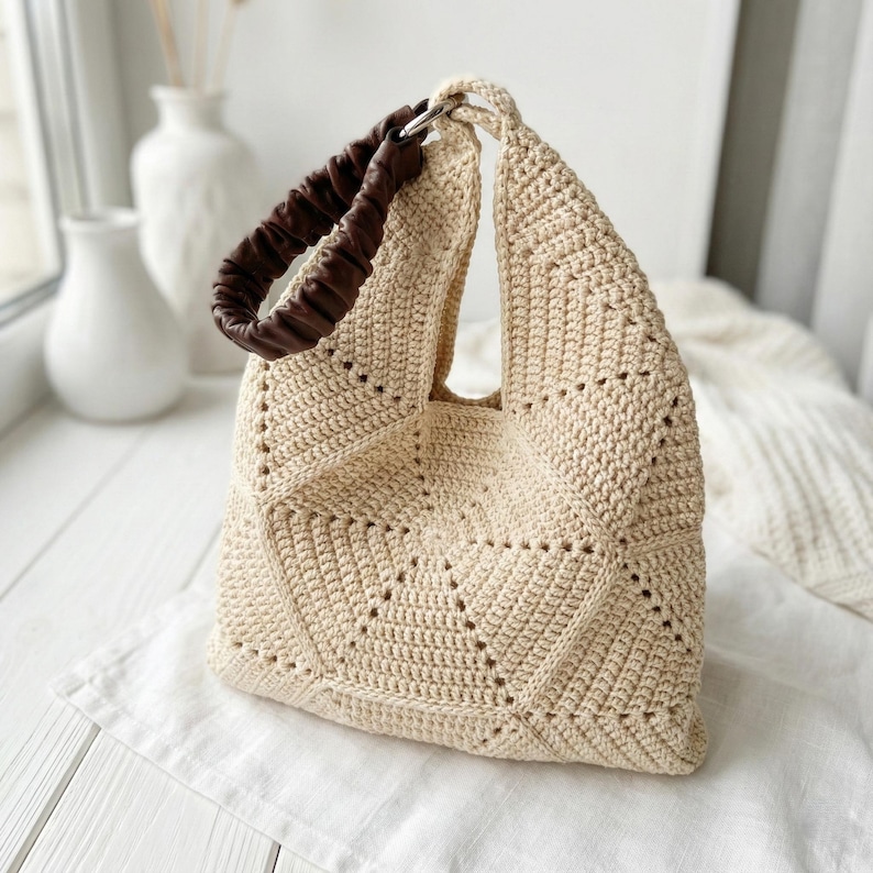 ALT TEXT (5)
1) Two-tone crochet tote bag in sage green and cream with a padded dark handle displayed on a light tabletop.
2) Side view of the patchwork crochet handbag showing triangle panels and a structured shape.
3) Close-up of the geometric triangle motif and textured stitches on the crochet tote bag.
4) Detail of the padded handle and hardware attachment at the top of the crochet bag.
5) Front view of the crochet tote highlighting the centered patchwork design and neat edging.