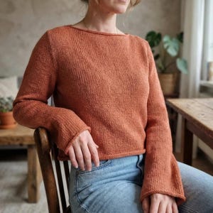 May include: A rust-colored knit sweater with a boat neck and long sleeves. The sweater has a ribbed texture and is paired with blue jeans. The sleeves have a flared design. The person is sitting on a wooden chair.