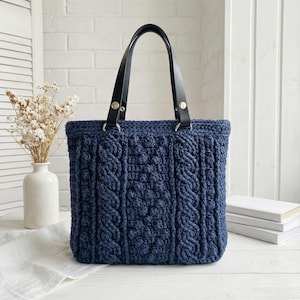 Puede incluir: Un bolso tote azul marino tejido a mano con un diseño de punto trenzado. El bolso tiene asas de cuero negro sujetas con herrajes plateados. El bolso es rectangular y está sobre una superficie blanca.