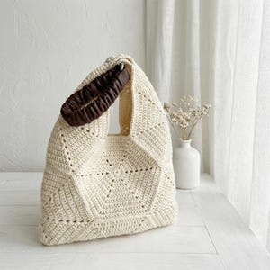 May include: Cream-coloured, crocheted tote bag with a geometric pattern. The bag has a brown leather handle. The bag is on a white surface. A white vase with dried flowers is in the background.