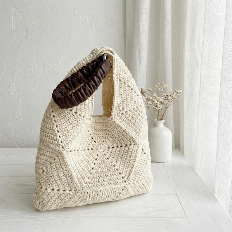 May include: Cream-coloured, crocheted tote bag with a geometric pattern. The bag has a brown leather handle. The bag is on a white surface. A white vase with dried flowers is in the background.