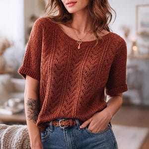 以下が含まれることがあります： テクスチャードニット模様の、錆色の半袖セーター。セーターはラウンドネックとロールカフス仕様。モデルはブルージーンズと茶色のベルトを着用。ペンダント付きのゴールドネックレスが見える。