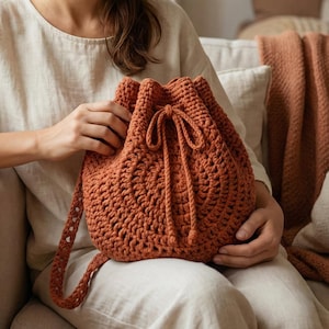 May include: A rust-colored, crocheted drawstring bag with a shoulder strap. The bag is made of thick yarn and features a textured pattern. The drawstring closure is tied with a matching cord. The bag is held by a person wearing a cream-colored shirt.