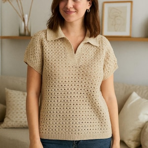 May include: A beige, short-sleeved polo shirt with a collared neckline. The shirt has a textured, open-knit design. The model is wearing blue jeans. The background includes a sofa, decorative pillows, and a framed picture.