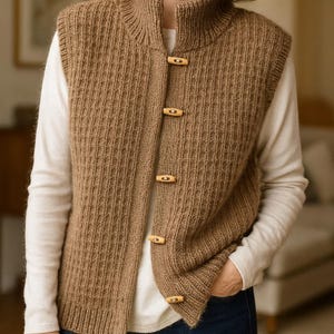 Peut inclure: Un gilet marron en tricot avec un col montant et des boutons en bois. Le gilet a une texture côtelée et est porté sur un t-shirt blanc à manches longues. Le modèle porte un jean bleu.