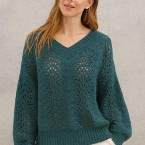 以下が含まれることがあります： 独特なオープンニットデザインの、ティールグリーンのVネックセーター。長袖とリブ編みの裾が特徴です。モデルは白いパンツを着用しています。カジュアルにもドレッシーにも着こなせる、スタイリッシュなセーターです。