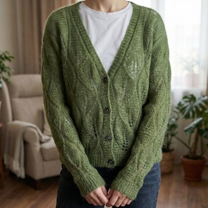 Puede incluir: Un cárdigan verde con botones y cuello en V. El suéter de punto tiene un patrón texturizado y se usa sobre una camiseta blanca. El cárdigan tiene mangas largas y está hecho de un material suave y difuso. El fondo incluye una silla, plantas y una ventana.