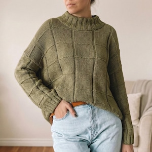 Op de afbeelding: Olijfgroene trui met een hoge halslijn en een ruitpatroon. De trui met lange mouwen is gemaakt van gebreid materiaal. De persoon draagt een lichtblauwe jeans en een bruine riem.