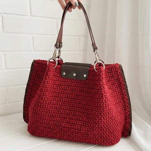 Peut inclure: Sac fourre-tout en crochet rouge fait à la main avec des poignées et des bordures en cuir marron. Le sac a une forme rectangulaire avec une fermeture à rabat et des accessoires argentés. Le sac est tenu par une main.