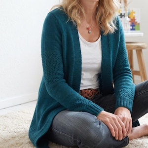 May include: A teal open-front cardigan featuring long sleeves with textured knit panels on the forearms and ribbed cuffs. The garment is worn over a plain white t-shirt paired with dark grey denim jeans and a brown leather belt with a woven buckle.