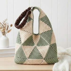 May include: A handmade crochet tote bag with a geometric pattern in cream and sage green. The bag features a brown, gathered leather handle and a large, open top. The bag is sitting on a wooden surface.