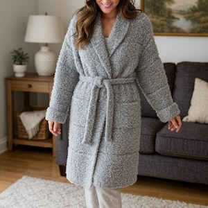 May include: A grey, long-sleeved dressing gown with a shawl collar and a matching tie belt. The robe has a textured, plush appearance and is knee-length. The person in the image is wearing light grey trousers. The background includes a sofa, rug, and side table.