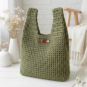 Puede incluir: Bolso tote de ganchillo verde oliva hecho a mano con asas resistentes. El bolso presenta una etiqueta de cuero marrón con una remache metálico. El bolso está hecho de un punto de tejido abierto texturizado.