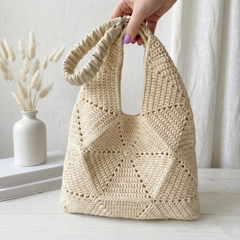 ALT TEXT (5)
1) Two-tone crochet tote bag in sage green and cream with a padded dark handle displayed on a light tabletop.
2) Side view of the patchwork crochet handbag showing triangle panels and a structured shape.
3) Close-up of the geometric triangle motif and textured stitches on the crochet tote bag.
4) Detail of the padded handle and hardware attachment at the top of the crochet bag.
5) Front view of the crochet tote highlighting the centered patchwork design and neat edging.