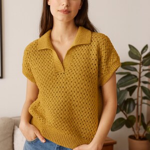 May include: Mustard yellow short-sleeved knit polo shirt with a collared neckline. The shirt has a textured, open-weave design and is paired with blue denim jeans. The model is standing indoors with hands in pockets.