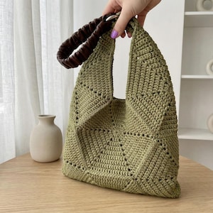 May include: An olive green crochet tote bag with a geometric design. It has a brown, gathered leather handle attached with silver-tone hardware. The bag is displayed on a wooden table. A neutral-toned vase is visible in the background.