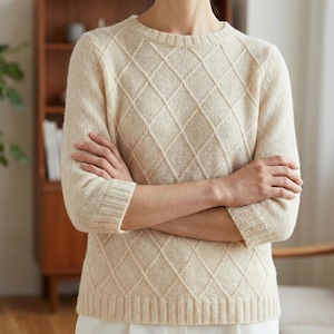 Peut inclure: Pull crème à motif losanges. Ce pull à manches longues a un col rond et des poignets et un ourlet côtelés. Les bras sont croisés. Le pull est fait d'un tricot doux et texturé.