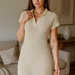 May include: A cream-colored, short-sleeved crochet dress with a collared neckline. The dress has a fitted silhouette and a textured, open-weave design. The model is standing in front of a neutral-toned background.