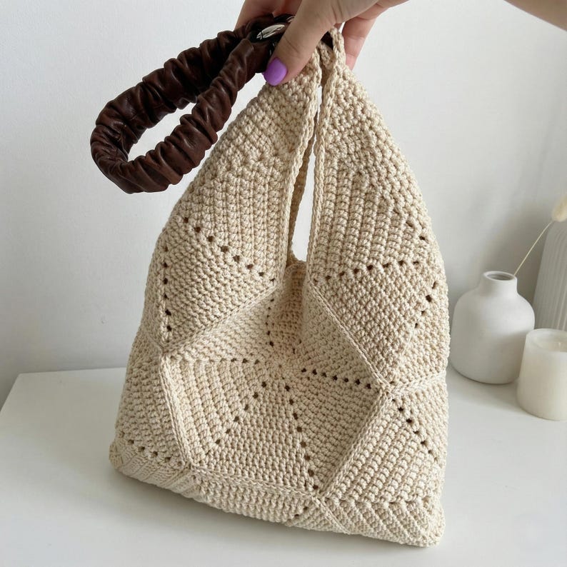 May include: Cream-coloured, handmade crochet tote bag with a geometric design. The bag features a brown, gathered leather handle. The bag is held up by a hand.